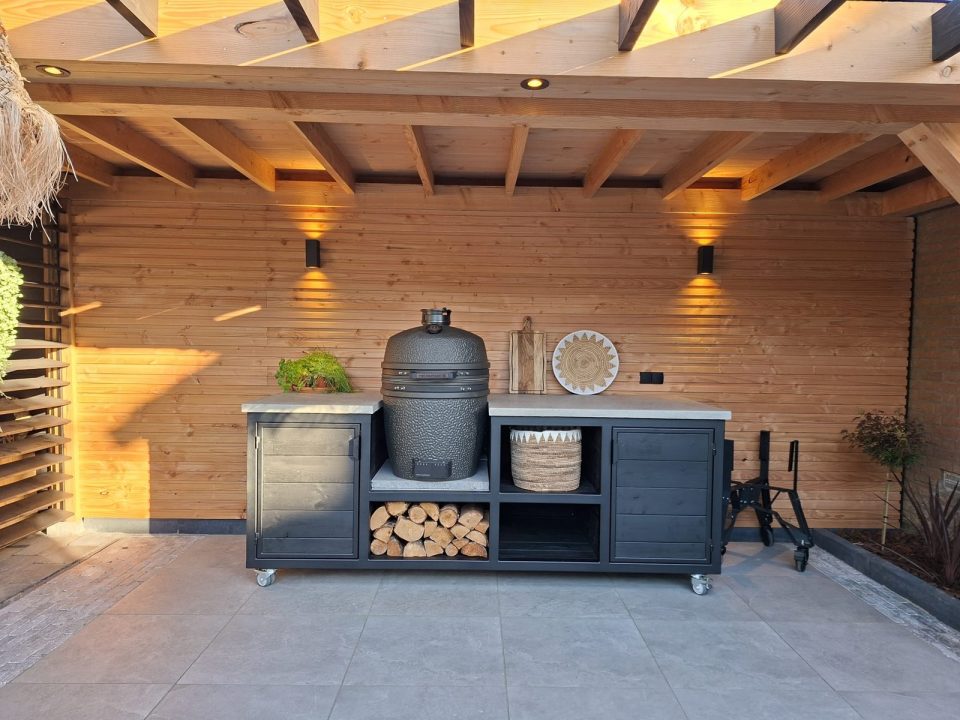 Zwarte buitenkeuken met kamado, licht werkblad, kasten en open opslagvakken onder houten overkapping